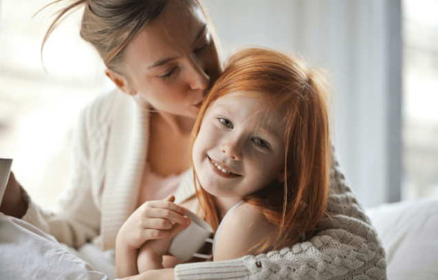 Mother and daughter sitting on a couch. the mother is kissing her daughter on hear head.