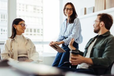 Three people in an office, man and two women. they are having a lively discussion about meidation. they are in a brightly lit, modern office space, next to a large window.