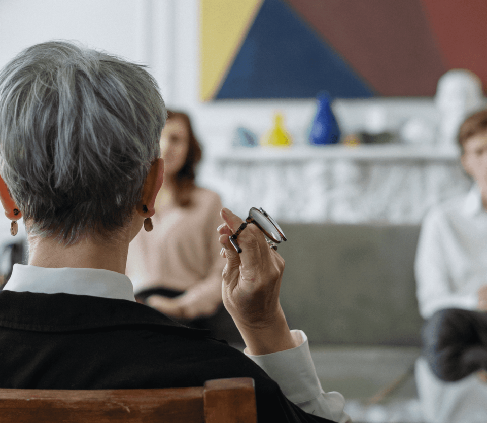 Older woman, and young couple sitting in the office. The older woman is listening intently to the couple. Older woman is facing away from the viewer and is in the foreground while the couple sit on a couch in the background.